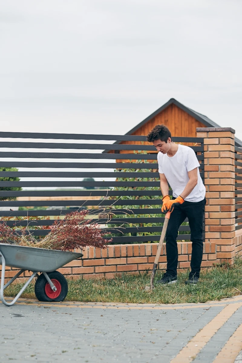 Robotnik usuwa chwasty łopatą obok ogrodzenia z bloczków z metalowymi przęsłami